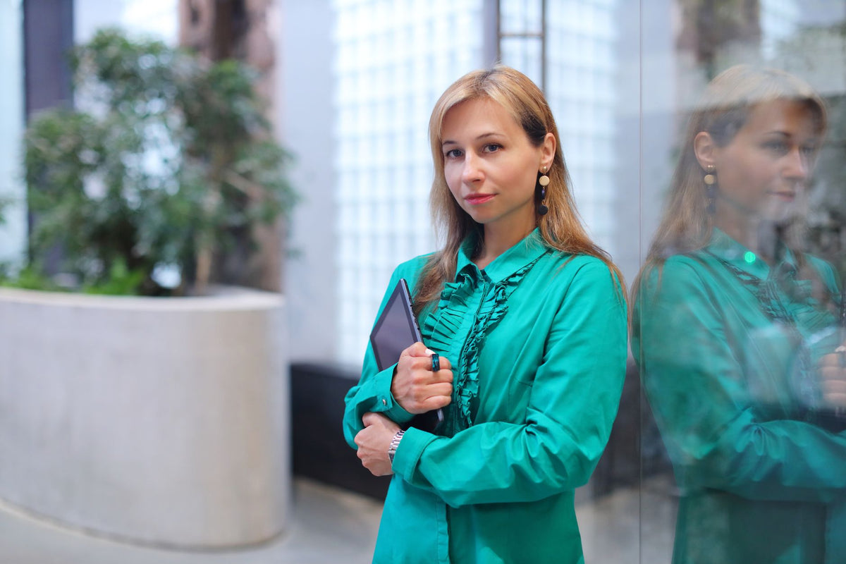 Elegant businesswoman with a tablet, confident and stylish in a turquoise blouse, leaning against a glass wall.