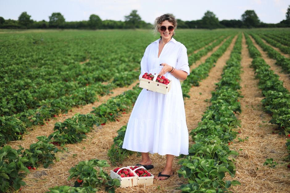 Woman in a white dress holding baskets of strawberries in a lush green field.