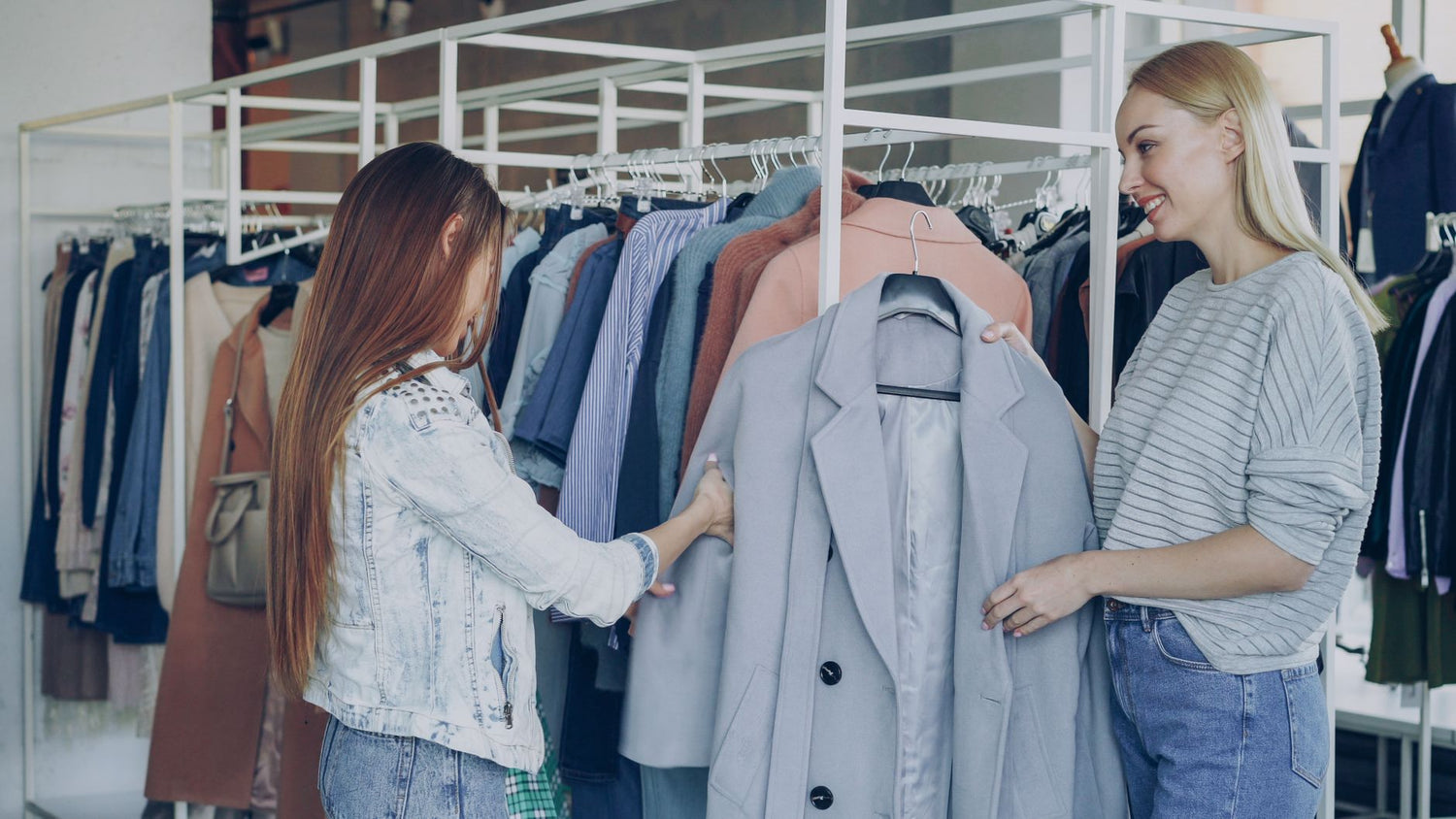 Two women shopping for jackets in a stylish clothing store, examining coats on a rack.