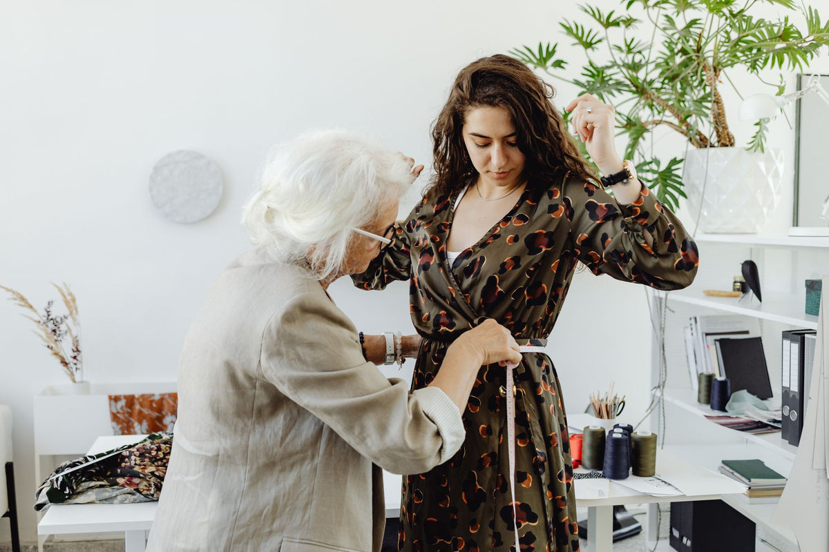 An elderly dressmaker adjusts a young woman's dress indoors, showcasing a personal fashion touch.