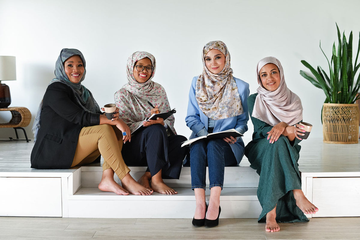 Four diverse women in hijabs discussing and working together indoors on wooden steps.