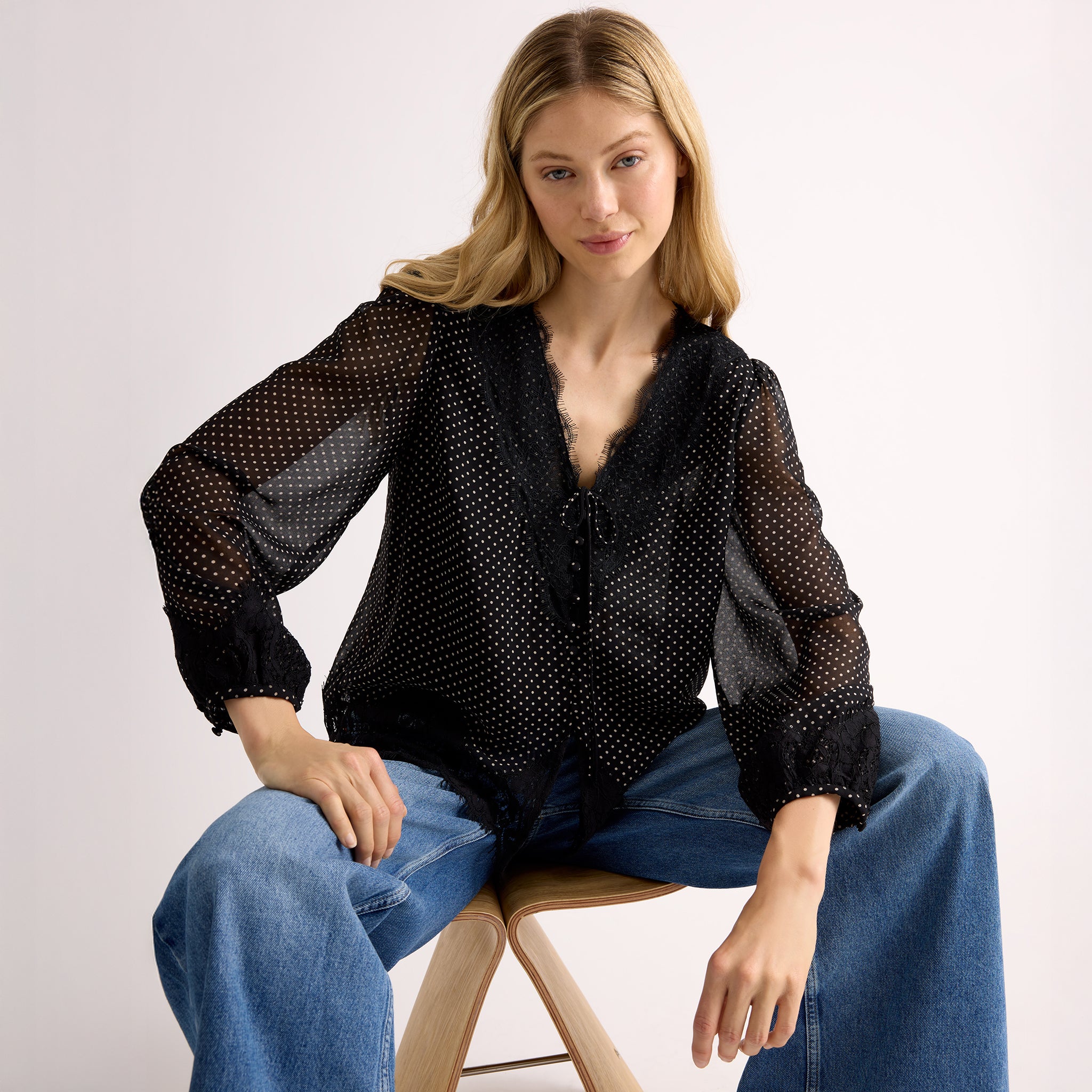 Woman wearing a black polka dot blouse and blue jeans sitting on a stool against a plain background